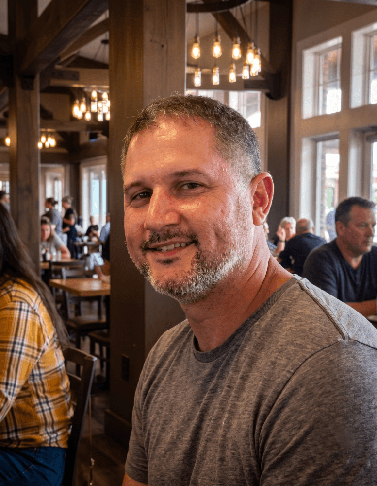 Man in a gray shirt smiling in a restaurant with wooden decor.