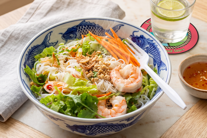 Bowl of noodle salad with shrimp, vegetables, and sauce, on a wooden tray next to a drink with lime.