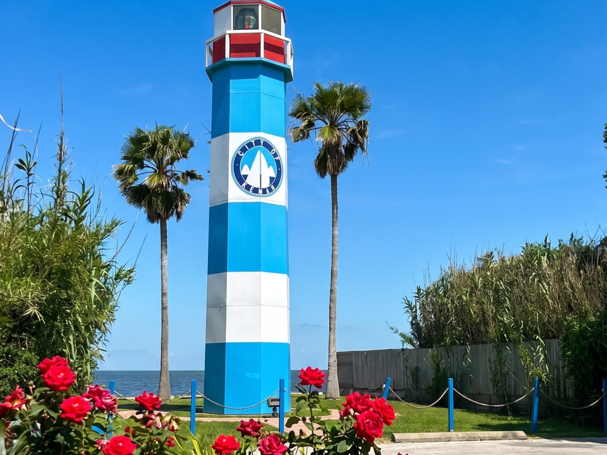 Blue and white lighthouse with red top, surrounded by flowers and palm trees.