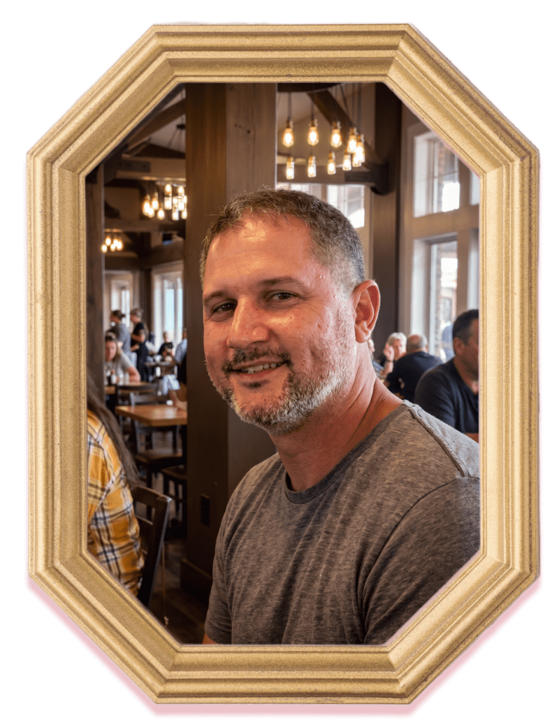 Man smiling in a wooden framed portrait, with people dining in the background.