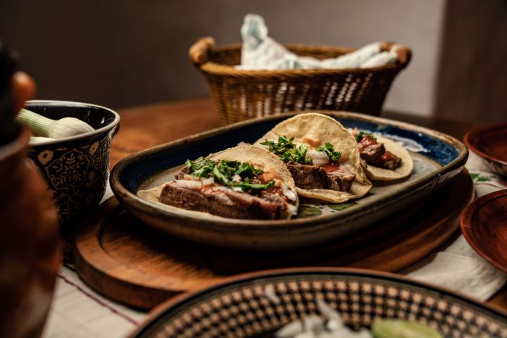 Plate with two tacos topped with beef and herbs, basket in background.