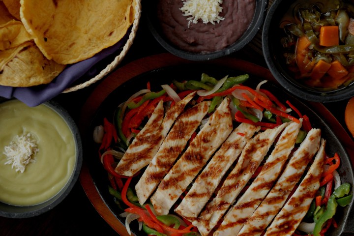 Grilled chicken strips with bell peppers, guacamole, chips, and sides in bowls.