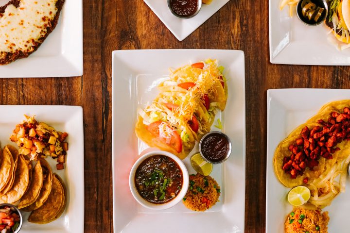 Various plated dishes with tacos, burgers, fries, and sides on a wooden table.