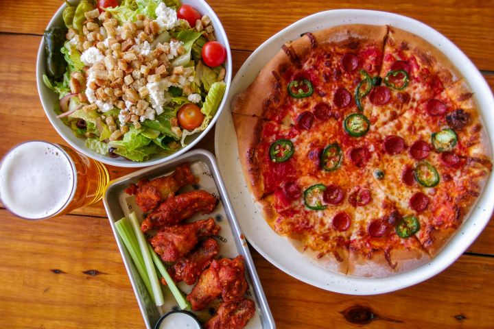 Pizza, salad, chicken wings, and a glass of beer on a wooden table.