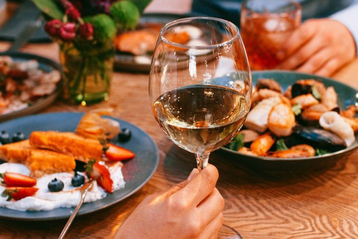 People dining with wine, plates of food, and flowers on a wooden table.