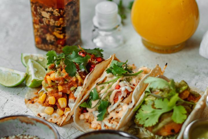 Tacos with toppings, sauces, and drinks on a gray surface.